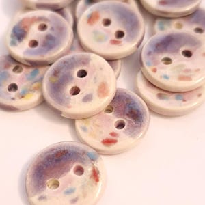 May include: A collection of ceramic buttons with two holes each. The buttons are round with a cream-colored base and feature a mottled design in shades of purple, pink, red, and blue. These decorative buttons are ideal for sewing and craft projects.