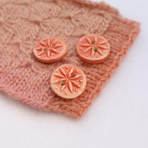 May include: Three round, peach-coloured ceramic buttons featuring a raised, embossed starburst or floral pattern with small dots around the perimeter. Each button has two central holes for attachment and is resting on a textured, peach-coloured knit fabric.