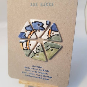 May include: A set of six handmade ceramic buttons in a triangular shape, each with unique abstract designs in blue, orange, and green. The buttons are attached to a tan card with the text "ZOE MAKES" and shop details.