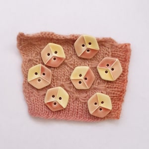 May include: Seven hexagonal buttons with a smooth, glossy finish, each featuring a two-tone color scheme of pale yellow and soft peach. The buttons have three holes arranged in a triangular pattern and are displayed on a textured, knitted fabric.