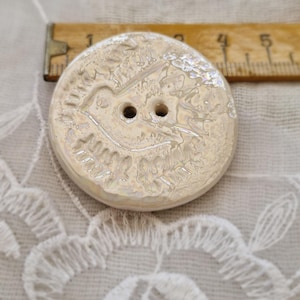 May include: A round, iridescent button with a bird design. The button has two holes and is approximately 1.5 inches in diameter. The button is made of a shiny, pearlescent material and has a textured surface.