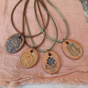 May include: Four ceramic pendants on brown and green cords. The pendants feature a hamsa hand, a sun, a flower, and a leaf.