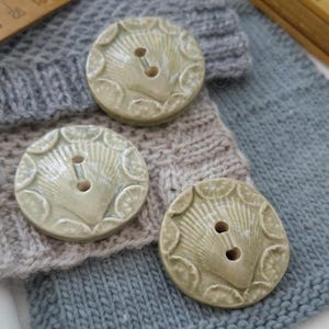 May include: Three round, light green ceramic buttons with a seashell design and two holes. The buttons are on a background of knitted fabric in shades of gray and blue, with a wooden ruler in the background.