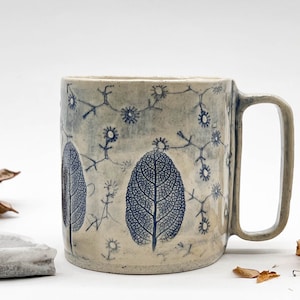 May include: A ceramic mug with a light beige base color and a rectangular handle. The mug is decorated with blue leaf and floral patterns. The leaves are detailed with intricate vein patterns. The mug is suitable for coffee, tea, or other beverages.