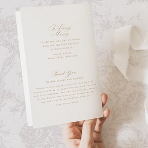 May include: A white card with gold lettering that reads "In Loving Memory" and includes a thank you message to family and friends. The card is being held by a hand.