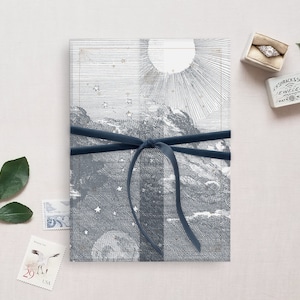 May include: A blue ribbon tied around a white and gray printed card with a celestial design. The card features a sun, stars, and mountains. The text "Ashrack & Sons Jewelers Watchmakers" is printed on a small white box.