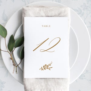 以下が含まれることがあります： 白いカードに金色の文字で「TABLE 12」と書いてあり、下部に小さな花柄のデザインが施されています。カードは、白いプレートの上に置かれたベージュのリネンナプキンの上に置かれています。