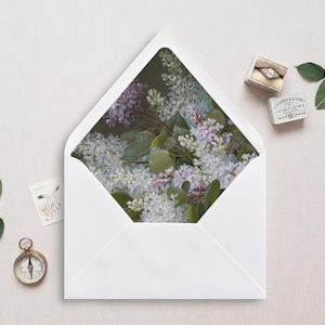 Pode incluir: Um envelope branco com um design floral na aba interna. O design apresenta um buquê de flores roxas e brancas com folhas verdes.