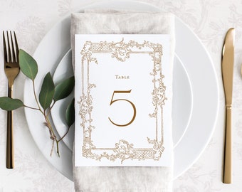 Könnte beinhalten: Eine weiße Karte mit einem goldenen, verzierten Rahmen und dem Text "Tisch 5" in goldenen Buchstaben. Die Karte liegt auf einer beigefarbenen Leinenserviette.