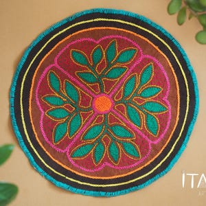 May include: Circular embroidered wall hanging with a colourful floral design in shades of pink, green, orange, and brown. The design features a central flower with four petals and leaves. The outer edge of the hanging is trimmed with a turquoise blue border.