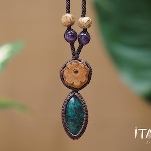 May include: A macrame necklace featuring a dark brown cord, two light tan speckled beads, two purple amethyst beads, a circular carved wooden flower pendant, and a marquise-shaped teal and black stone pendant. The text ITAYA ARTE is visible.