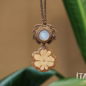 May include: A handmade macrame necklace featuring a light blue stone pendant and a unique, flower-shaped pendant made of wood and resin. The necklace is suspended from a brown cord. The background is a blurred mix of green and brown.