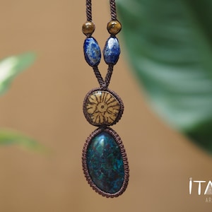 May include: A macrame necklace featuring a dark brown cord, two small round beads, two blue lapis lazuli stones, a central circular tan pendant with a floral pattern, and a large oval dark teal stone pendant. The text ITAYA ARTE is visible.