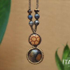 May include: A macrame necklace featuring a tan cord with two circular pendants. The top pendant is a light tan disc with brown spots, and the bottom pendant is a dark brown and tan stone. The cord includes blue and grey beads and the text ITAYA ARTE.