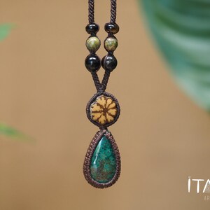 May include: A macrame necklace featuring a teardrop-shaped green stone pendant, a circular tan floral-patterned bead, and smaller dark brown and green accent beads on a brown cord. The text ITAYA ARTE is visible in the bottom right corner.