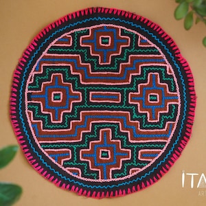 May include: A round, black, embroidered wall hanging with a geometric pattern in pink, green, and blue. The pattern is symmetrical and features a central square with four smaller squares around it. The wall hanging is surrounded by a pink and black border.