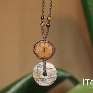 May include: A handcrafted macrame necklace featuring a flower-shaped wooden pendant and a circular stone pendant. The necklace has a brown cord with decorative beads. The logo "ITAYA ARTE" is visible in the bottom right corner.