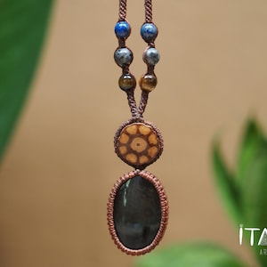 May include: A macrame necklace featuring a brown cord, a large oval dark gray stone pendant, and a circular wooden accent piece. The cord is decorated with smaller blue, gray, and brown beads. The text ITAYA ARTE is visible in the bottom right corner.