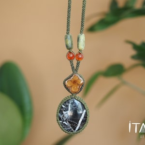 May include: A green cord necklace with a black and white stone pendant, a brown flower pendant, and green and orange beads. The necklace is hanging against a light brown background with green foliage.
