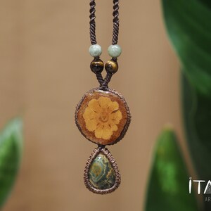 May include: A macrame necklace featuring a brown cord, two light green beads, two brown tiger eye beads, a circular wooden pendant with a carved flower design, and a teardrop-shaped green and brown stone pendant. The text ITAYA ARTE is visible.