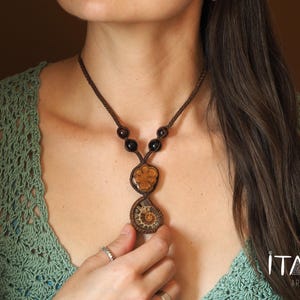 Può includere: Una collana di corda marrone con un ciondolo di ammonite marrone e dorato e perline marroni. La collana è indossata da una persona con i capelli lunghi e castani.