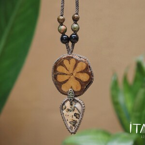 May include: A macrame necklace featuring a circular wooden pendant with a six-petaled flower design, suspended above a teardrop-shaped stone pendant with brown and cream speckles. The cord includes dark brown and green beads near the top.