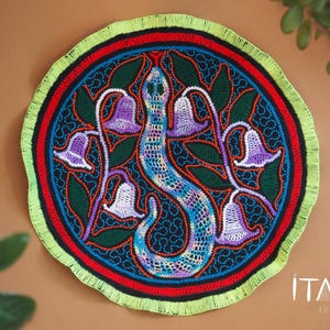 Può includere: Un arazzo da parete circolare colorato con un motivo di serpente verde, circondato da fiori viola e foglie verdi. Lo sfondo è blu scuro con bordi rossi e gialli. Il testo "ITAYA ARTE" è visibile nell'angolo in basso a destra.
