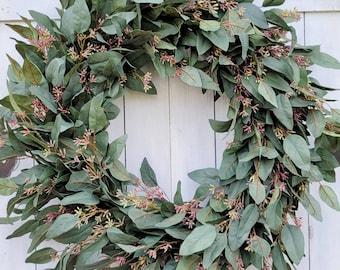 May include: A green eucalyptus wreath with small pink flowers. The wreath is made of artificial leaves and flowers and is perfect for decorating a door or wall.