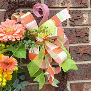 Op de afbeelding: Een krans met een roze, groene en oranje gestreepte strik. De krans is versierd met perzikkleurige bloemen en groene bladeren.