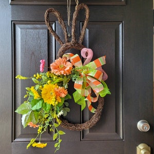 Op de afbeelding: Een lentekrans met oranje en gele bloemen, groene bladeren en een roze en oranje gestreept lint. De krans is gevormd als een konijn en hangt aan een bruine deur.