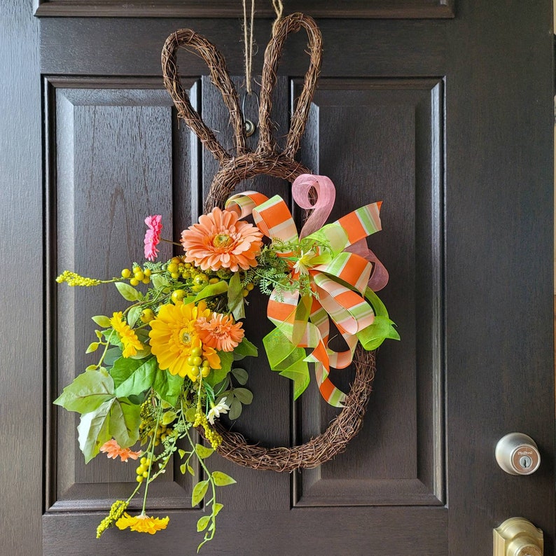 Op de afbeelding: Een lentekrans met oranje en gele bloemen, groene bladeren en een roze en oranje gestreept lint. De krans is gevormd als een konijn en hangt aan een bruine deur.