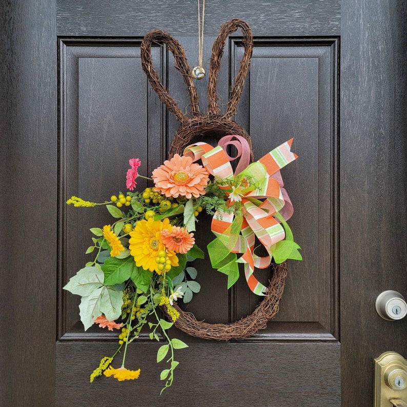 Op de afbeelding: Een bruine krans in de vorm van een konijn met verschillende bloemen, waaronder roze, gele en oranje bloemen, en een groen-wit gestreept lint.