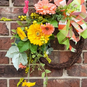Op de afbeelding: Een bruine takkenkrans met een groen-oranje gestreepte strik. De krans is versierd met roze, gele en groene bloemen.