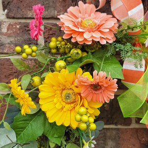 Op de afbeelding: Een zomerse krans met roze, oranje en gele bloemen. De krans is versierd met groene bladeren en kleine gele bessen.