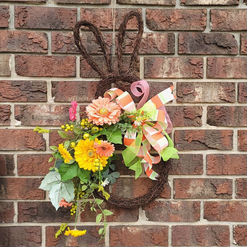 Op de afbeelding: Een bruine takkenkrans in de vorm van een konijn met een roze, groen en wit lint en een boeket gele en perzikkleurige bloemen.