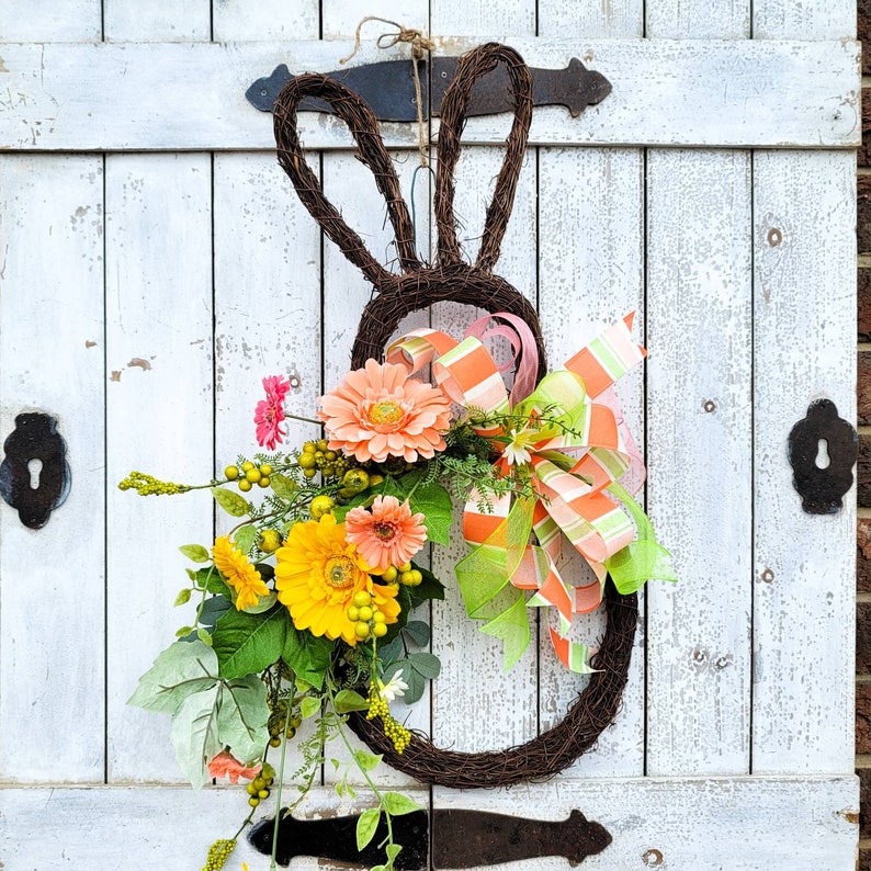 Op de afbeelding: Een bruine takkenkrans in de vorm van een konijn met een roze en groen lint en een boeket gele en roze bloemen.