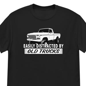 May include: Black t-shirt featuring a white graphic of an old pickup truck. The text "EASILY DISTRACTED BY OLD TRUCKS" is printed below the truck in a bold, white font. The shirt is made of a soft, comfortable material.