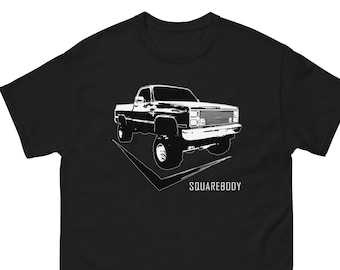 Peut inclure: T-shirt noir avec une silhouette blanche d'un pick-up classique à carrosserie carrée. Le camion est de face et le texte "SQUAREBODY" est imprimé sous le camion.