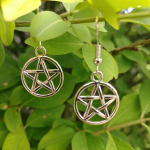 May include: Silver-colored pentagram earrings hanging from silver hooks. The earrings are circular with a five-pointed star design. The background is a lush green with leaves and branches.