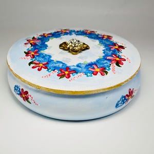 May include: A round, light blue decorative box with a floral design. The lid features a wreath of red flowers and blue accents, with a gold-colored jewel-encrusted clasp. The box has a gold trim.