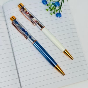 May include: Two ballpoint pens with rose gold accents and clear barrels filled with small tumbled gemstones. One pen has a metallic blue lower body, while the other has a white lower body. Both feature rose gold clips and conical tips.