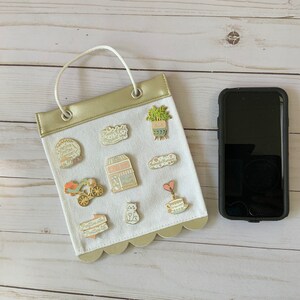 May include: A white canvas bag with a gold top and scalloped edges. The bag has a handle and is decorated with various enamel pins. The pins feature images of a coffee shop, a plant, a cat, a teacup, a heart, and a bicycle. The bag is hanging on a white wooden surface.