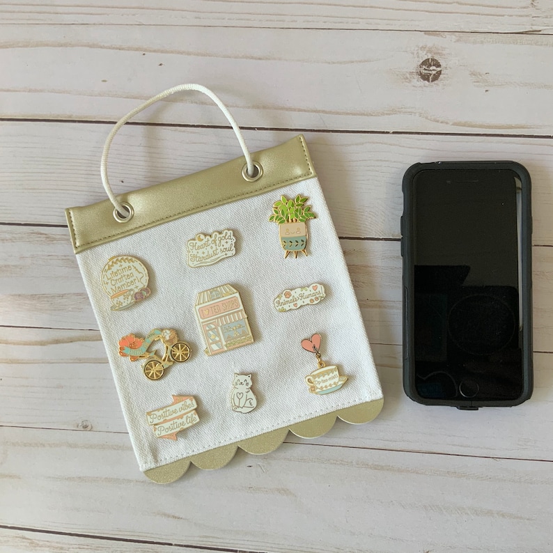 May include: A white canvas bag with a gold top and scalloped edges. The bag has a handle and is decorated with various enamel pins. The pins feature images of a coffee shop, a plant, a cat, a teacup, a heart, and a bicycle. The bag is hanging on a white wooden surface.