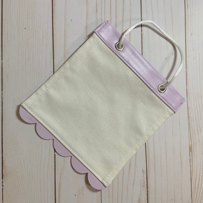 May include: A small, white canvas bag with a lavender scalloped edge and a lavender faux leather top. The bag has two metal eyelets and a white rope handle.