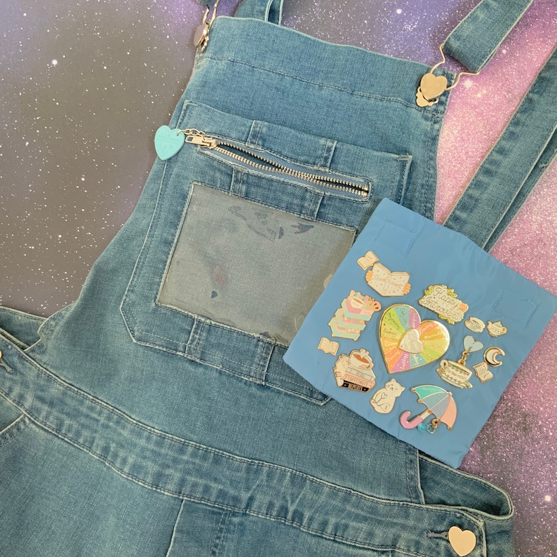 May include: A pair of blue denim overalls with a zippered pocket and a clear plastic window. The overalls have a light blue heart-shaped charm attached to the zipper. A blue piece of felt with a variety of gold enamel pins is attached to the overalls.
