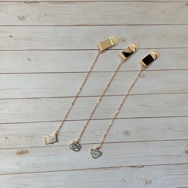 May include: Three gold bookmark chains with different charms. The charms are a book, a teacup, and a teapot. The chains are hanging from a wooden surface.