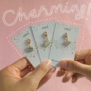 Könnte beinhalten: Drei goldfarbene Charms mit weißen und hellblauen Emaille-Designs werden auf weißen Karten präsentiert. Die Charms zeigen eine Teetasse, ein offenes Buch und eine Teekanne. Das Wort "Charming!" steht in Schreibschrift darüber.