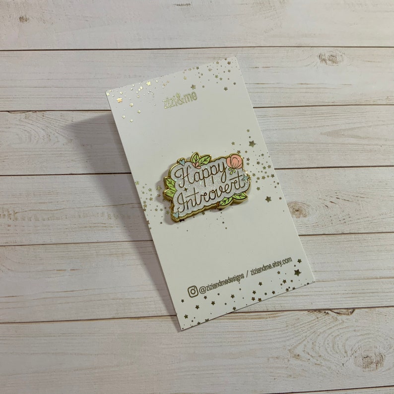 May include: Gold enamel pin with a white background and green and pink floral accents. The pin says "Happy Introvert" in a cursive font.