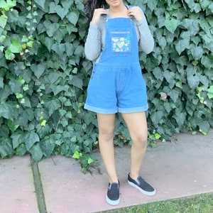 May include: A woman wearing a blue denim overall with a clear plastic pocket on the front. The pocket has colorful patches and designs. She is wearing black slip-on shoes.
