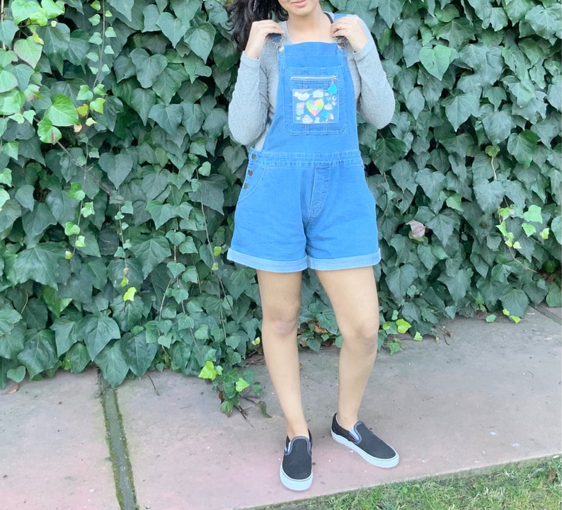 May include: A woman wearing a blue denim overall with a clear plastic pocket on the front. The pocket has colorful patches and designs. She is wearing black slip-on shoes.